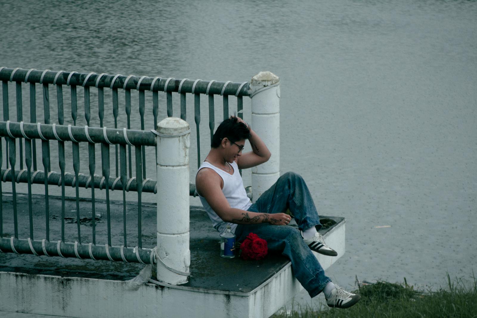 Young man in casual attire sits by river, immersed in thought, capturing serene urban ambiance.